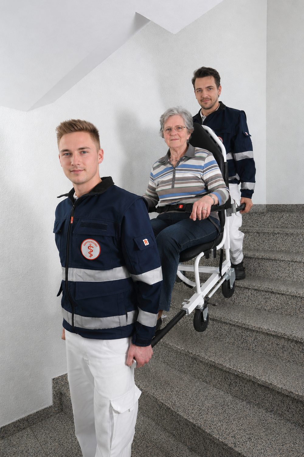 Zwei Medimobil-Mitarbeiter tragen eine Patientin im Tragestuhl über eine Treppe